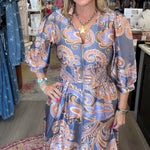 A woman modeling a long-sleeve midi dress with a blue, orange, and white paisley pattern inside a boutique.
