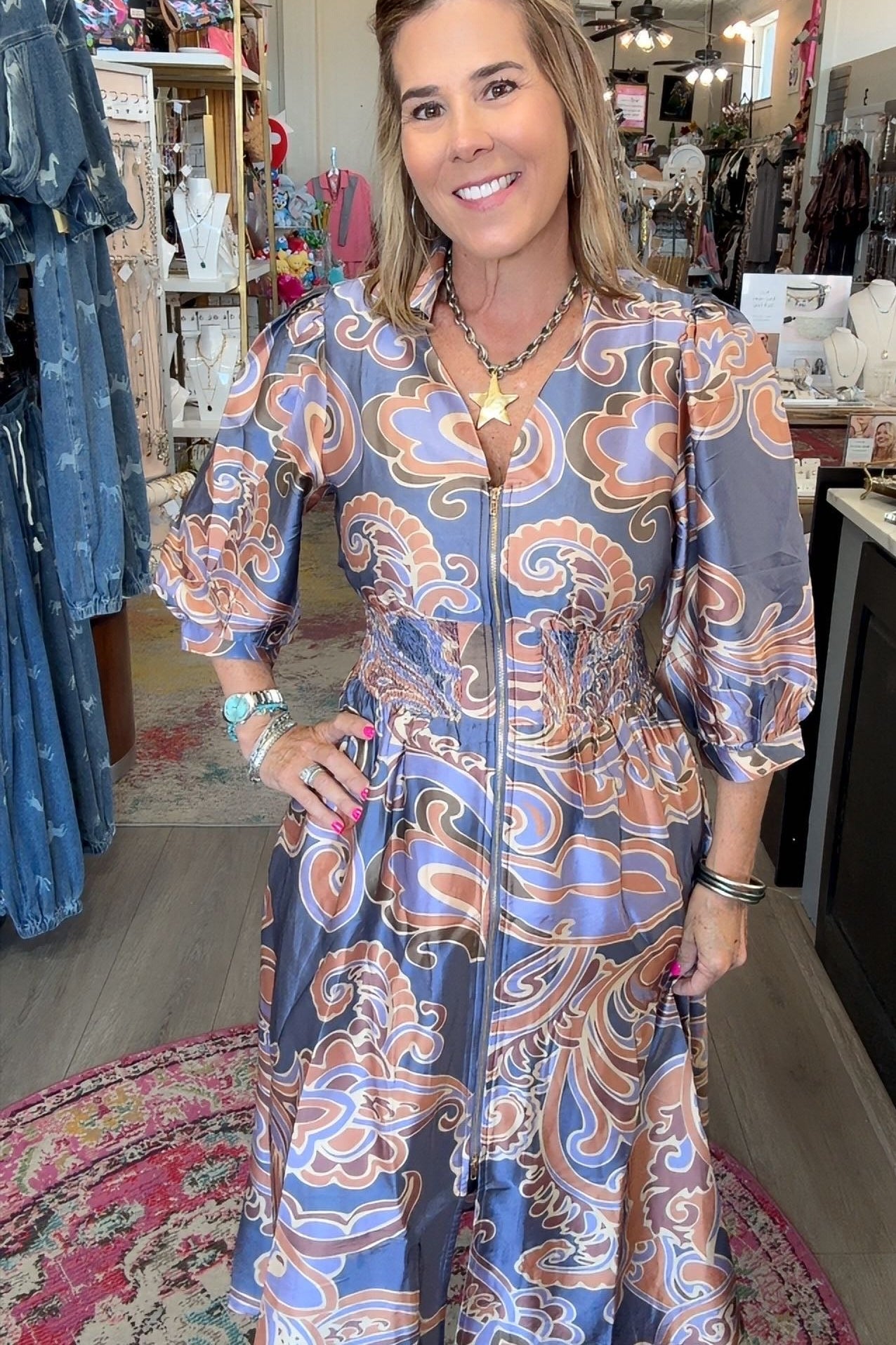 A woman modeling a long-sleeve midi dress with a blue, orange, and white paisley pattern inside a boutique.