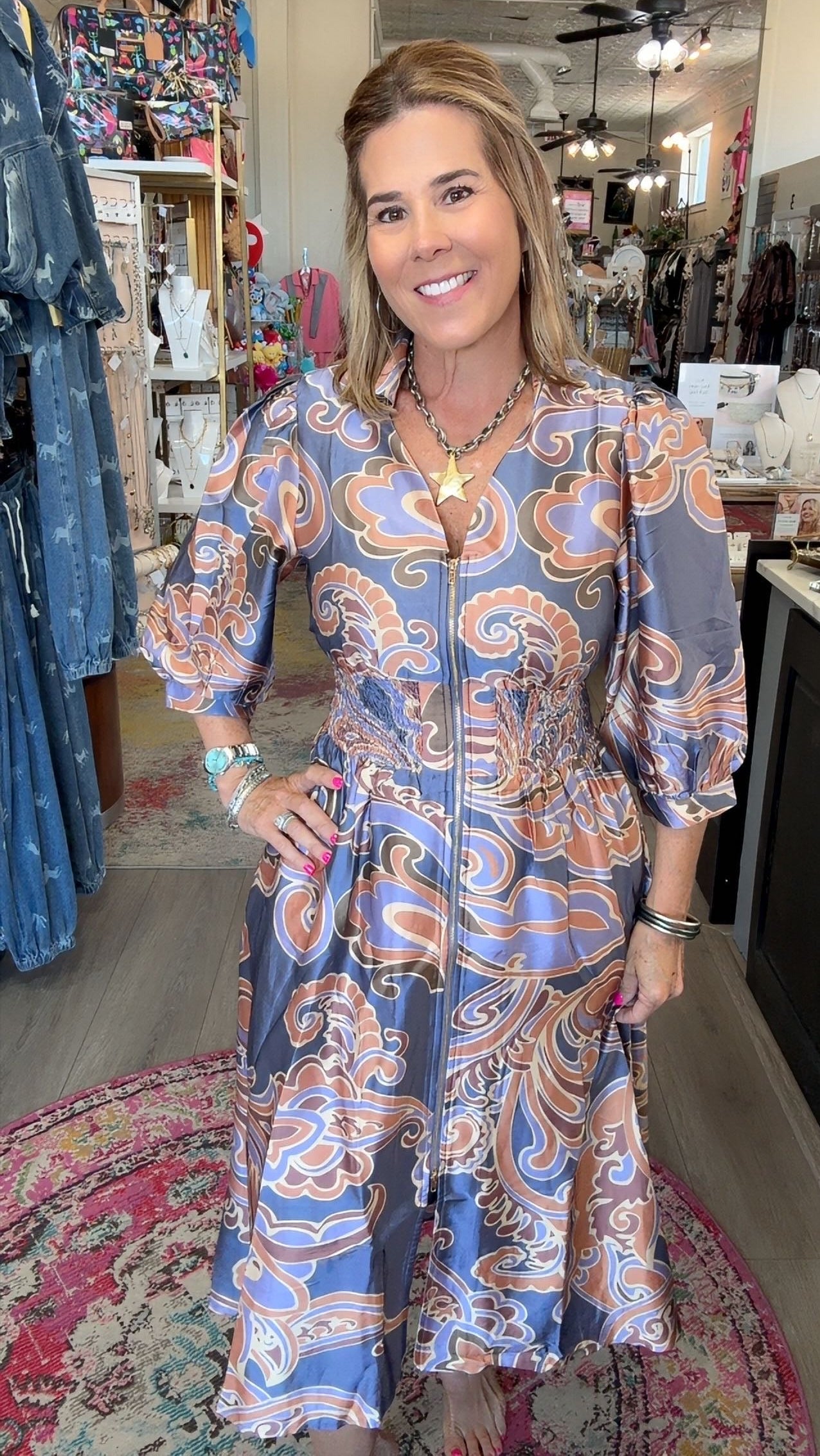 A woman modeling a long-sleeve midi dress with a blue, orange, and white paisley pattern inside a boutique.