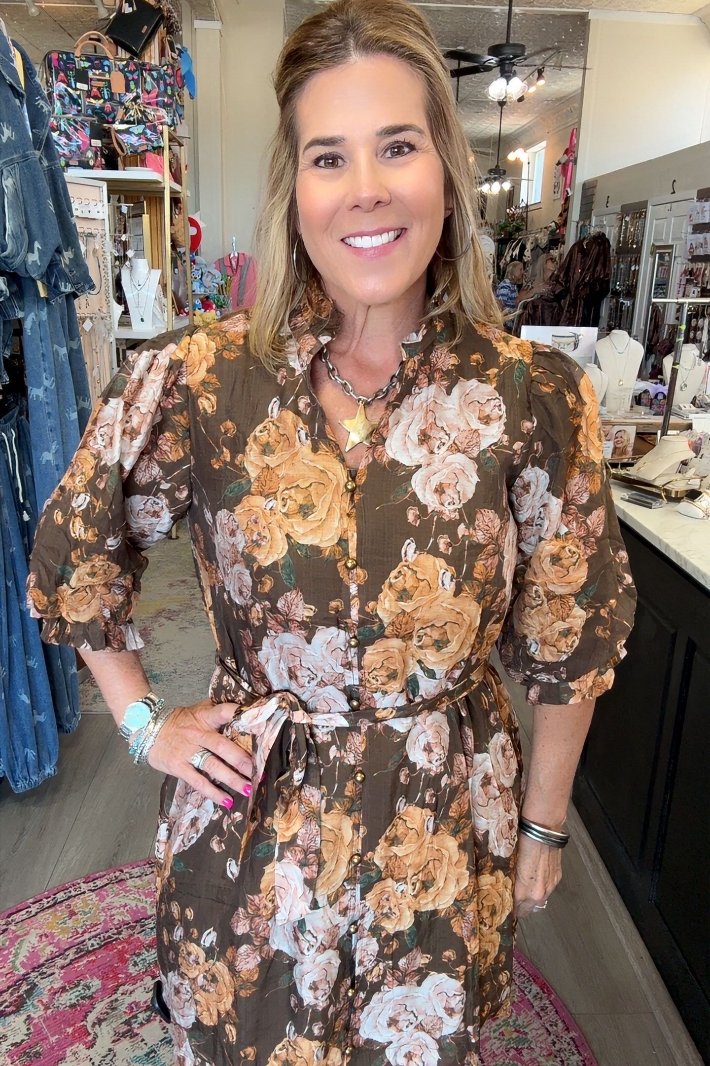 A woman modeling a brown short-sleeve floral romper with beige, white, and orange flowers, standing inside a boutique.