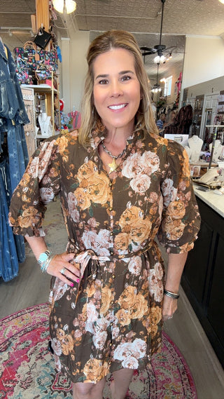 A woman modeling a brown short-sleeve floral romper with beige, white, and orange flowers, standing inside a boutique.