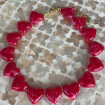 A heart-shaped beaded necklace with glossy red heart charms and a gold-tone clasp, displayed on a textured mosaic tile surface.