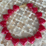 A heart-shaped beaded necklace with glossy red heart charms and a gold-tone clasp, displayed on a textured mosaic tile surface.