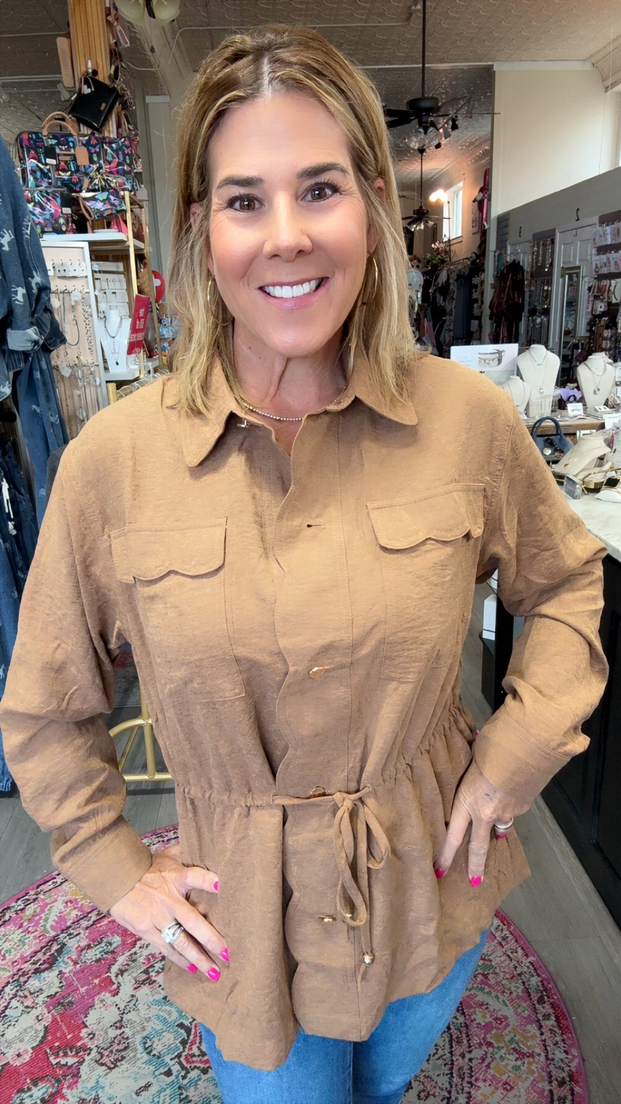 A woman modeling a tan long-sleeve button-up shirt with chest pockets and a drawstring waist, standing inside a retail store.