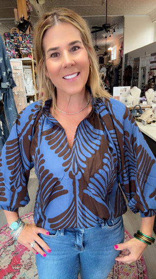 A woman wearing a blue and brown palm-leaf patterned blouse with blue jeans, standing in a retail store.