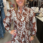 A woman modeling a brown and white botanical print long-sleeve shirt dress inside a boutique store.