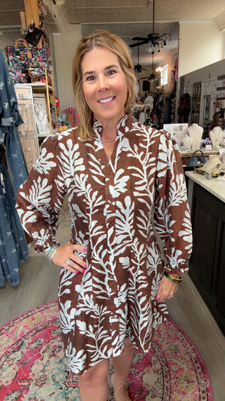 A woman modeling a brown and white botanical print long-sleeve shirt dress inside a boutique store.