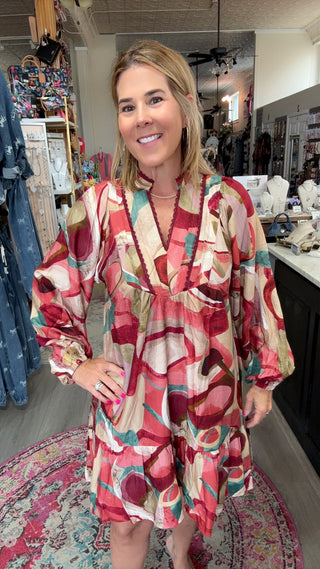 A woman modeling a long-sleeve, V-neck mini dress with a colorful abstract pattern in red, pink, beige, and teal, standing inside a boutique.