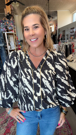 A woman with blonde hair stands in a boutique wearing a black and cream abstract patterned blouse with gold trim, paired with blue jeans.