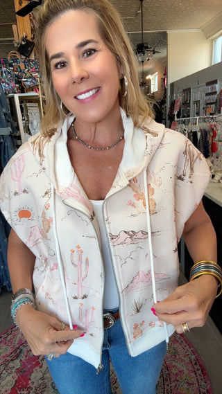 A woman wearing a sleeveless zip-up hoodie vest with a desert landscape print, standing inside a retail store.