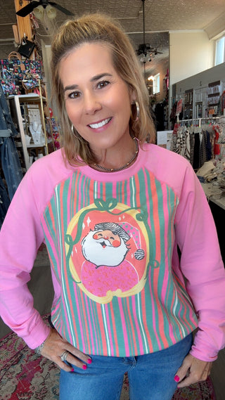 A woman wearing a pink long-sleeve sweatshirt with a colorful striped front panel and a cartoon Santa graphic, standing inside a clothing store.