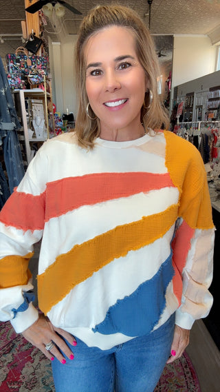 A woman wearing a colorful striped crewneck sweater with diagonal bands of white, orange, yellow, and blue, standing in a retail store.