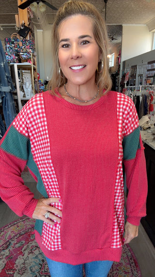 A woman wearing a long-sleeve crewneck sweater with a vibrant pink ribbed body, red and white gingham patterned side panels, and dark green gingham sleeve sections, standing in a retail store.
