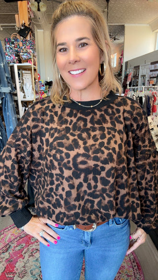 A woman wearing a brown and black leopard print long-sleeve top with black ribbed cuffs and collar, paired with blue jeans, standing inside a clothing store.