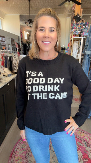 A woman wearing a black crewneck sweater with white text that reads 'IT'S A GOOD DAY TO DRINK AND THE GAME' in a retail store setting.