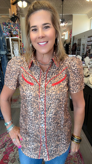 A woman modeling a short-sleeve leopard print button-down shirt with red western-style trim and chest pockets, paired with blue jeans in a retail store setting.