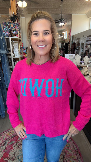 A woman wearing a bright pink knit sweater with the word 'HOW' in large turquoise letters across the chest, paired with blue jeans, standing inside a retail store.