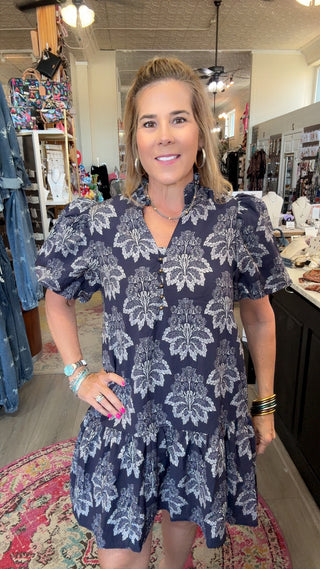 A woman modeling a navy blue short-sleeve dress with a white floral damask pattern, standing inside a boutique shop.