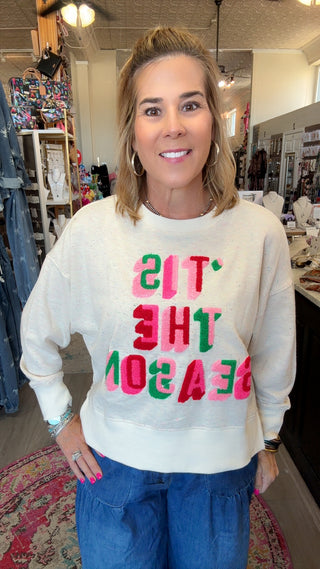 A woman wearing a cream-colored crewneck sweatshirt with colorful, hand-painted style text reading 'IT'S THE SEASON' in pink, green, and red, paired with blue jeans, standing inside a retail store.