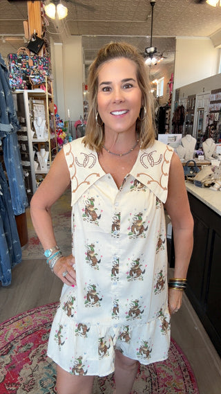 A woman modeling a sleeveless white romper with a brown and green cowboy print and embroidered details on the collar, standing inside a retail boutique.