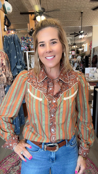 A woman modeling a long-sleeve western-style button-up shirt with vertical orange, green, and white stripes and a brown paisley bandolier collar, paired with blue jeans in a retail store setting.