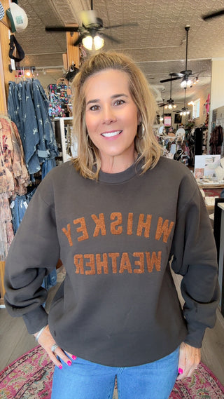 A woman smiling in a boutique shop, wearing a dark charcoal gray sweatshirt with 'WHISKEY WEATHER' printed in orange across the front, paired with blue jeans.