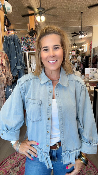 A woman smiling in a boutique while wearing a light-wash blue denim jacket with ruffled trim and balloon sleeves, paired with a white top and blue jeans.