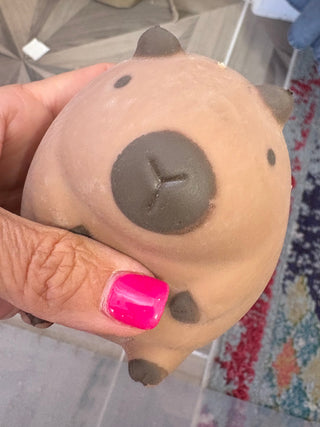 A hand with bright pink nail polish holds a small, squishy capybara toy. The toy is light tan with a dark brown snout, ears, and feet.