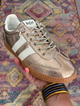 A metallic bronze leather sneaker with white side stripes and a gum sole, held by a hand against a colorful patterned rug background.