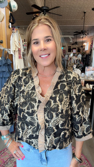 A woman with blonde hair stands in a boutique wearing a black and beige floral patterned blouse with ruffle trim and light blue jeans.
