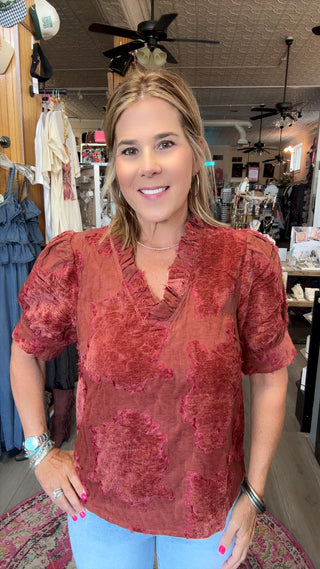 A woman modeling a rust-red short-sleeve blouse with a textured, tonal floral pattern, paired with light blue jeans in a boutique setting.