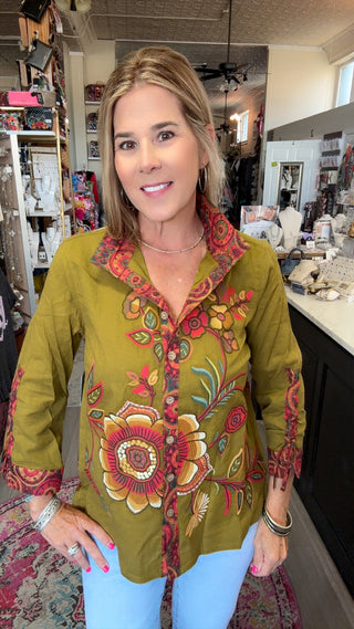 A woman wearing a vibrant olive green button-up blouse with colorful floral and paisley patterns, paired with light blue jeans, standing inside a retail boutique.