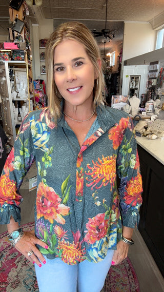 A woman wearing a long-sleeve button-up blouse with a vibrant multicolor floral print on a slate blue-gray background, paired with light blue jeans, standing inside a retail shop.