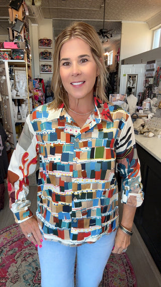 A woman modeling a long-sleeve button-up shirt with a colorful geometric patchwork pattern, standing inside a retail store.