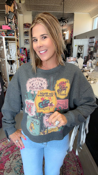 A woman wearing a dark gray graphic sweatshirt with a collage of vintage-style patches and text, paired with light blue jeans, standing in a retail store interior.