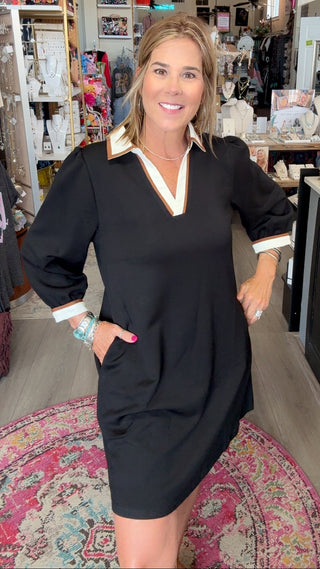 A woman modeling a black mini dress with a V-neckline, gold trim, and white striped cuffs in a retail store setting.