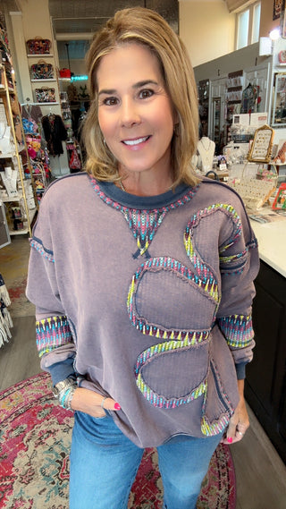 A woman wearing a lavender-gray long-sleeve sweatshirt with colorful geometric tribal-style embroidery, standing inside a retail store.