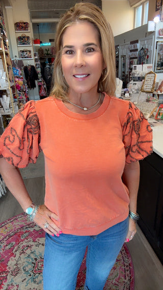 A woman wearing a coral orange short-sleeve top with patterned puff sleeves and blue jeans, standing inside a retail store.