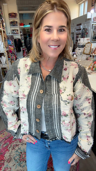 A woman wearing a patchwork quilted jacket with floral and striped panels, blue jeans, and a white top, standing inside a retail store.