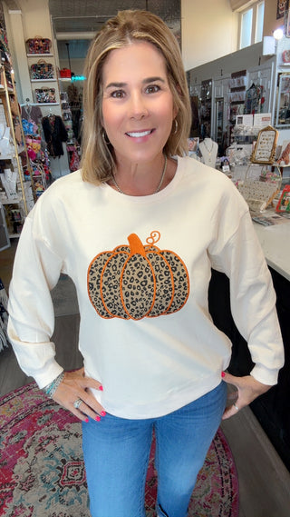 A woman wearing a cream-colored crewneck sweatshirt with a leopard-print pumpkin graphic on the front, paired with blue jeans, standing inside a retail store.