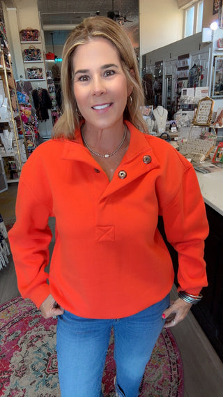 A woman wearing a bright orange long-sleeve polo-style top with blue jeans, standing inside a retail store.