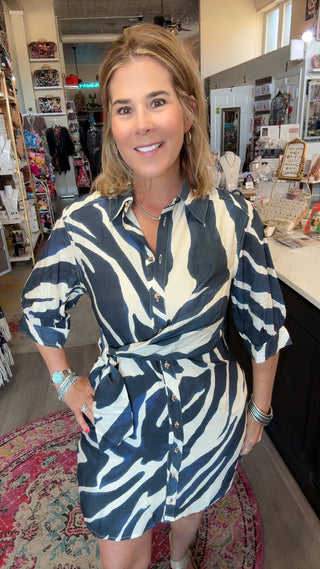 A woman wearing a navy blue and cream zebra-print shirt dress with a tie waist, standing in a retail boutique.