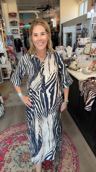 A woman wearing a black and white zebra-print midi shirt dress stands inside a boutique shop. The dress features a bold vertical zebra stripe pattern in black and off-white.
