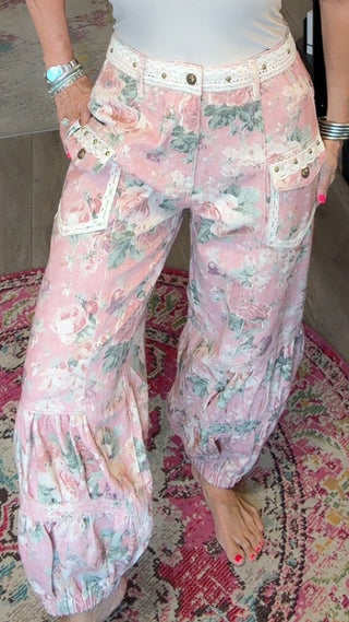 A woman stands barefoot inside a boutique shop, wearing a white ribbed tank top and light pink wide-leg floral pants with white pocket trim.