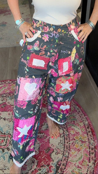 A woman stands in a retail store wearing floral patchwork pajama pants with a white tank top. The pants feature a dark background with pink, red, and white floral and star patterns.