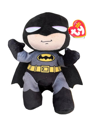 A Ty Beanie Baby plush toy of Batman, featuring a black and gray body with a yellow utility belt and chest emblem, resting on a colorful patterned carpet. A hand is holding the red Ty brand hangtag next to the plush.