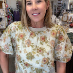A woman wearing a cream-colored short-sleeve blouse with a vintage floral pattern in green, orange, and pink, paired with blue jeans, standing inside a retail store.