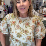 A woman wearing a cream-colored short-sleeve blouse with a vintage floral pattern in green, orange, and pink, paired with blue jeans, standing inside a retail store.