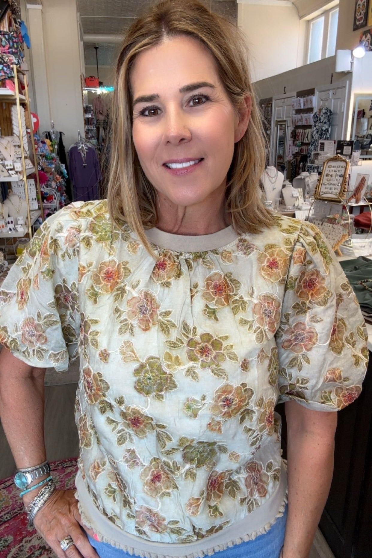 A woman wearing a cream-colored short-sleeve blouse with a vintage floral pattern in green, orange, and pink, paired with blue jeans, standing inside a retail store.
