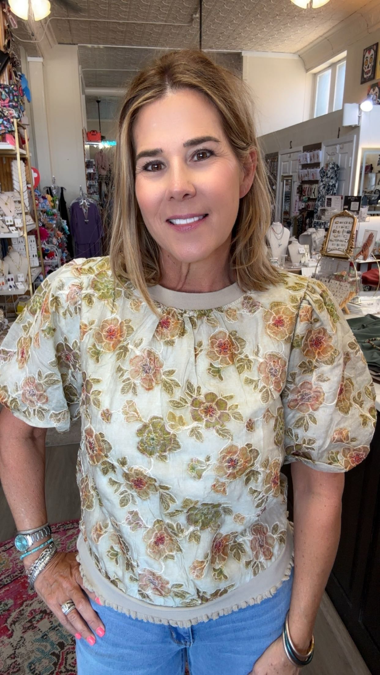 A woman wearing a cream-colored short-sleeve blouse with a vintage floral pattern in green, orange, and pink, paired with blue jeans, standing inside a retail store.